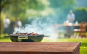 A wooden table in the foreground with a smoking barbecue grill and blurred people in the background outdoors, perfect for gathering and sharing accessible grilling tips.