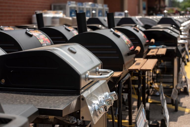 A row of outdoor gas grills lined up for sale outside a store, each with closed lids, control knobs, and wooden side shelves—showcasing options for those searching for the Best Time to Buy a BBQ Grill.