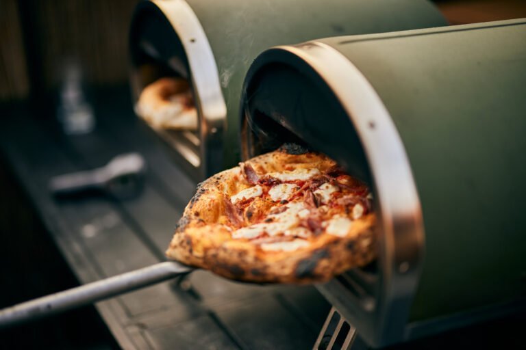 A freshly baked pizza is being removed from a small outdoor pizza oven using a metal peel, showcasing the convenience and quality offered by Best Value Pizza Ovens.