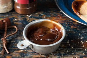 A small metal cup filled with thick barbecue sauce sits on a worn wooden table near a plate and spice containers, prompting the question: do you need to refrigerate BBQ sauce after opening?.