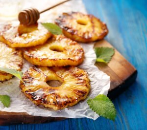 Several slices of grilled pineapple topped with honey and garnished with mint leaves are arranged on parchment paper over a wooden board, offering a tasty reminder of how BBQ can promote sustainability through fresh, locally sourced produce.
