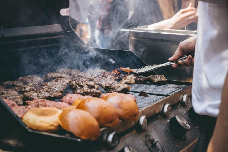 Several hamburger patties and buns are being grilled on a barbecue, with a person using a spatula to cook the Game-Day Grub. Smoke is rising from the grill.