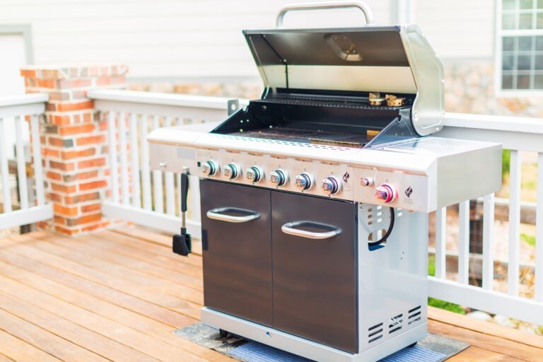 A stainless steel gas grill with six burners and an open lid sits on a wooden deck, with a cleaning brush hanging on the side. Wondering how heavy is a grill like this? Its sturdy build ensures lasting performance for outdoor cooking.