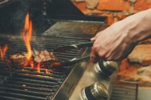 A hand uses tongs to grill steaks over an open flame on a gas barbecue, avoiding common mistakes in grilling & smoking meat, with a brick wall background.