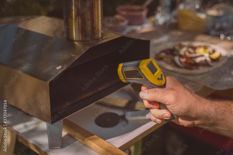 A hand holds an infrared thermometer—one of the most innovative BBQ gadgets—aimed at the opening of a metal pizza oven, with a pizza and various items visible in the background.