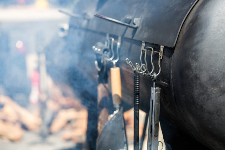 Barbecue tools with wooden handles hang on hooks in front of a smoker grill, smoke curling up and blurred logs behind—reminding you that maintaining your BBQ tools is key for top grilling performance.