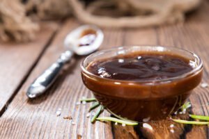 A small glass bowl filled with dark barbecue sauce sits on a wooden table, alongside a spoon and scattered rosemary and salt—perfect for gauging how much BBQ sauce per person you’ll need for your next gathering.