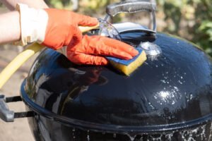 A person wearing orange gloves uses a sponge and hose to clean the lid of a black barbecue grill outdoors, demonstrating techniques similar to how to get BBQ grease off concrete.