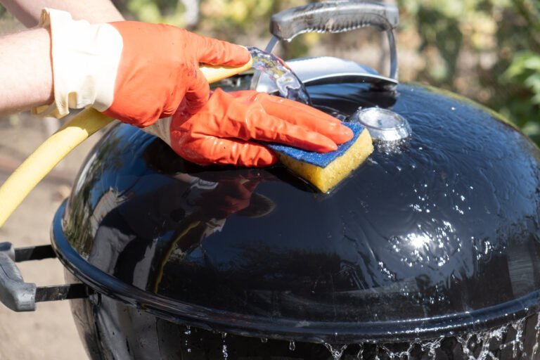 A person wearing orange gloves uses a sponge and hose to clean the lid of a black barbecue grill outdoors, demonstrating techniques similar to how to get BBQ grease off concrete.
