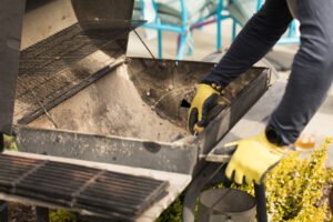 Wearing yellow gloves, a person cleans an open outdoor grill, removing ash and debris from the bottom section while considering what to do with BBQ ashes afterward.