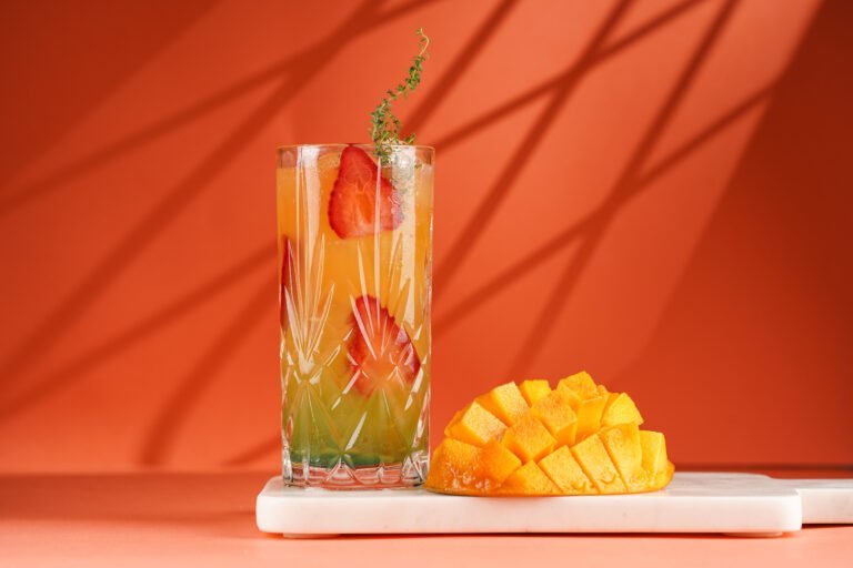 A tall glass of layered fruit drink with ice, garnished with strawberries and a sprig of herb, sits next to sliced mango on a white serving board—perfect inspiration for your summer cocktail recipes against an orange background.